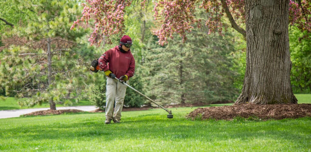 A Scott's Lawn Care technician trimming around landscaping and a tree.